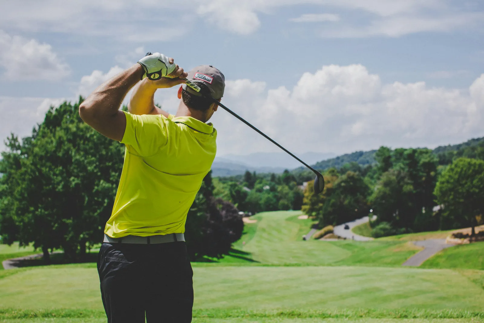 Golfer in a yellow shirt finishing a swing on a tree-lined course beneath a cloudy blue sky.