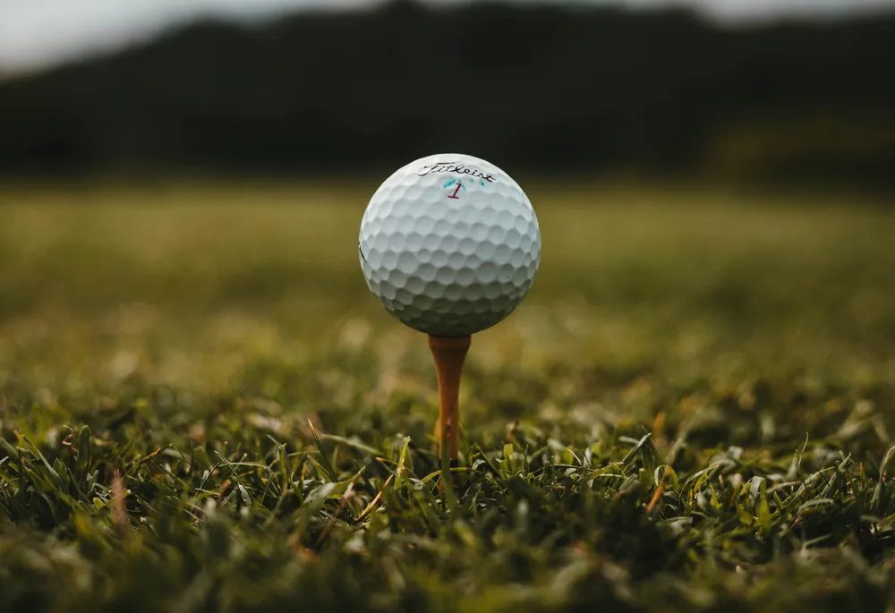 White golf ball resting on an orange tee in grass, with trees blurred behind it.